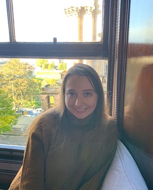 Photo of Molly sitting with a green jumper and trees in the background.