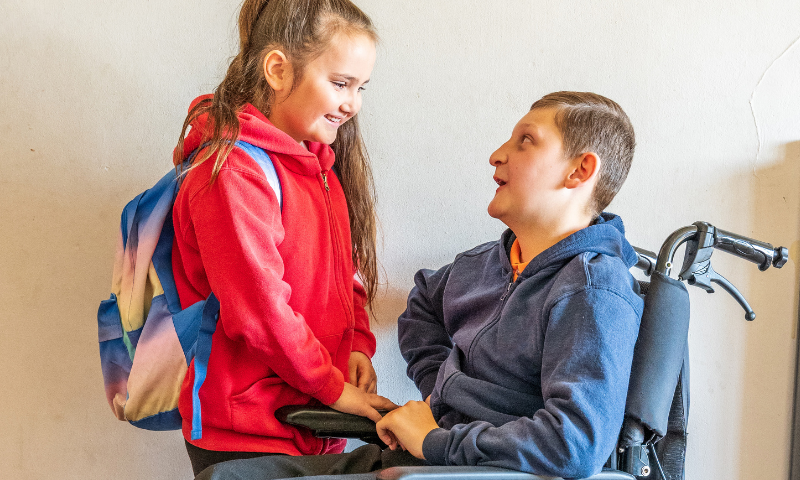 Photo shows a child holding hands with a family member who is a wheelchair user. They are smiling warmly at each other.