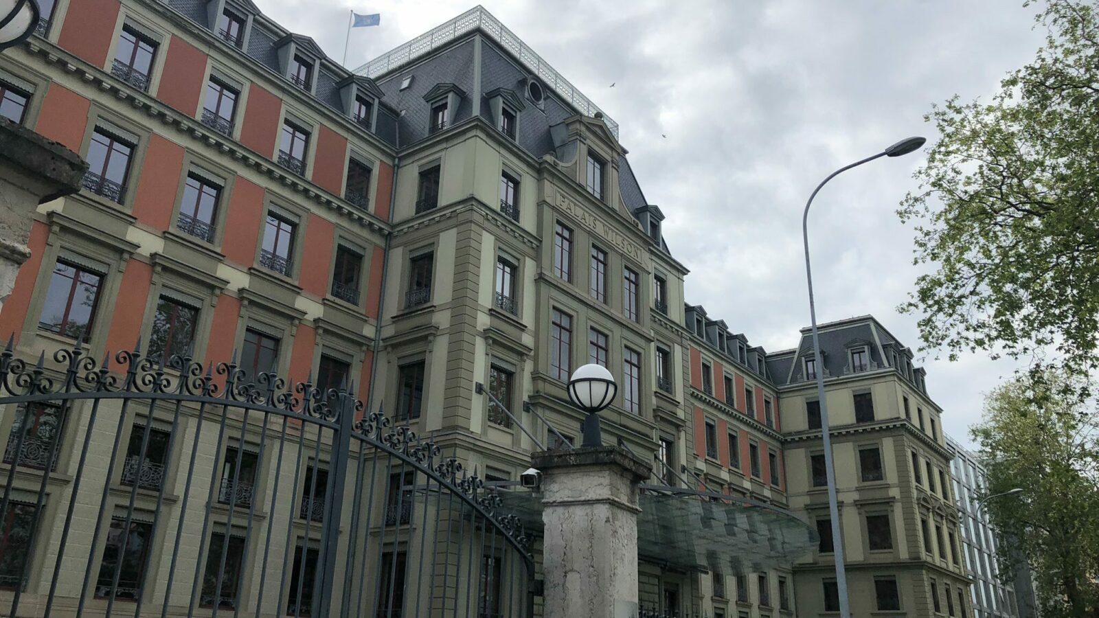 Photo shows Palais Wilson in Geneva where the UN Committee held the UK's child rights review. It is a tall, ornate building with orange and cream walls. It has lots of windows, pillars and a big security gate.