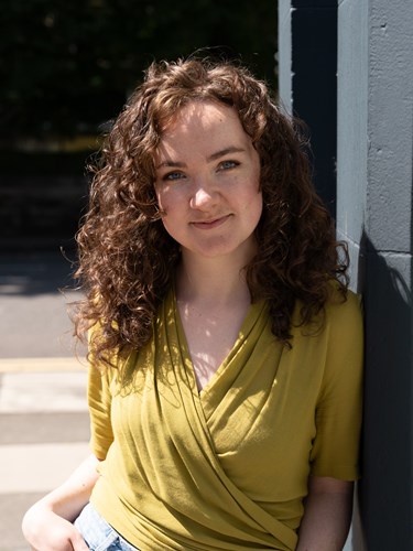 Photo of Maria, Legal and Policy Manager, green top on, leaning against a wall.