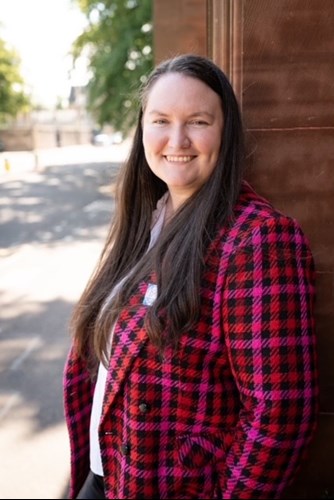 Photo of Sarah, Development Manager, leaning against a wall wearing a pink and red blazer.