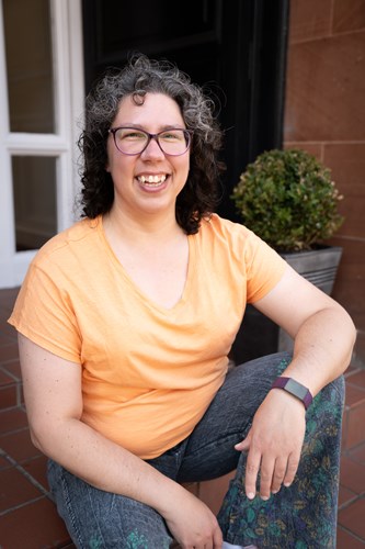 Photo of Christine, sitting on stairs wearing orange top with a door and plant pot in the background.