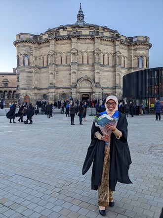 Photo of Siti standing in graduation gown in front of a building, smiling holding a bunch of flowers.